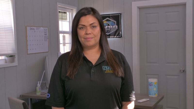 Woman standing in a gray office, wearing a black polo shirt, with a slight smile.