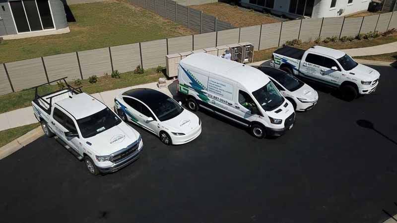 Five white utility vehicles parked in a row on a dark lot beside a fenced building.