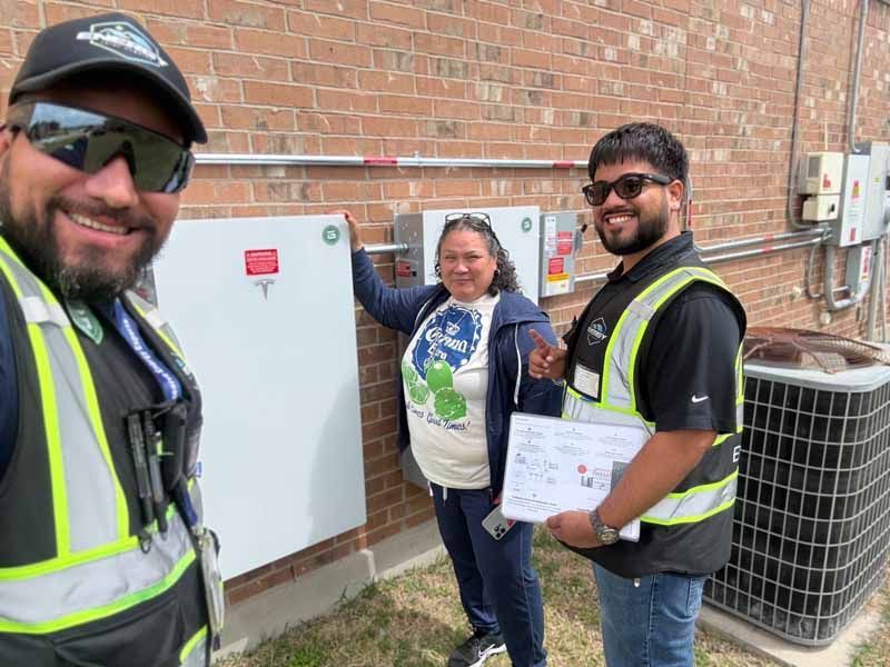 Three workers in safety vests stand by outdoor electrical panels against a brick wall.