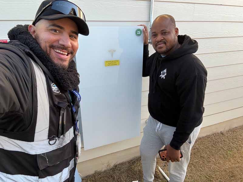Two men standing outside by a utility box, one smiling in a cap and vest, the other in a black hoodie and gray pants