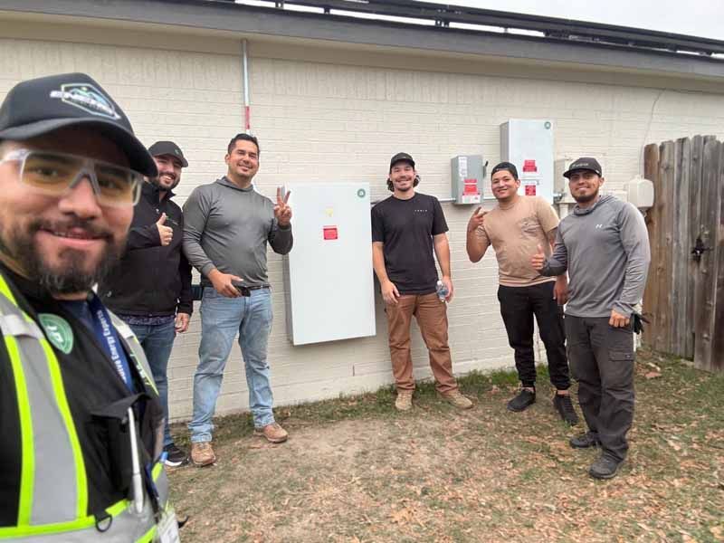 Five workers posing outdoors beside a wall-mounted electrical panel, one giving a thumbs-up