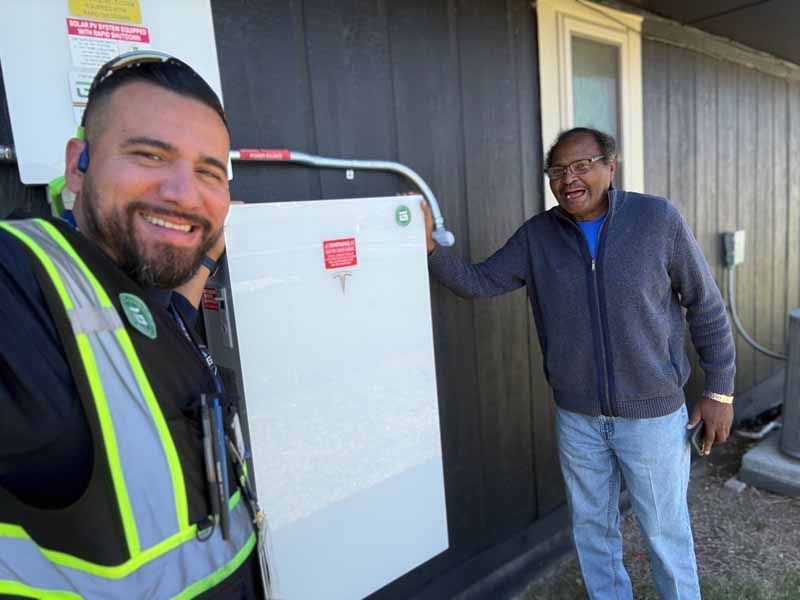 Two men standing beside a large white utility box outside a building, one in a reflective vest smiling at the camera.