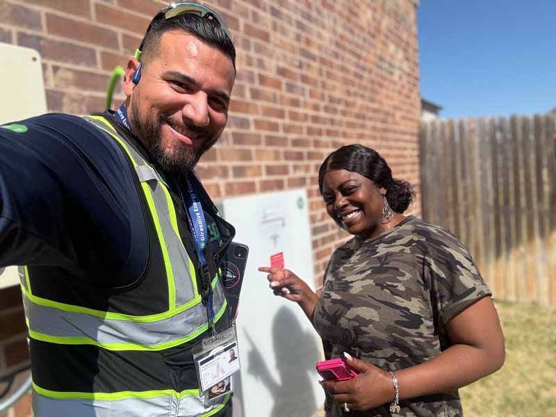 Two smiling people beside an electrical panel outdoors, one holding a key fob and one in a reflective safety vest