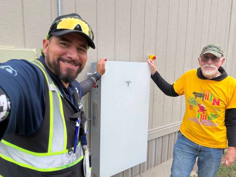 Two workers pose beside a white metal panel outdoors, one in a reflective vest and one in a yellow shirt.