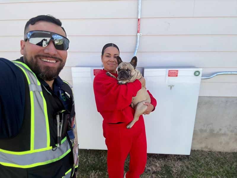 Two workers in safety gear posing outside a building, one holding a small dog in red coveralls.