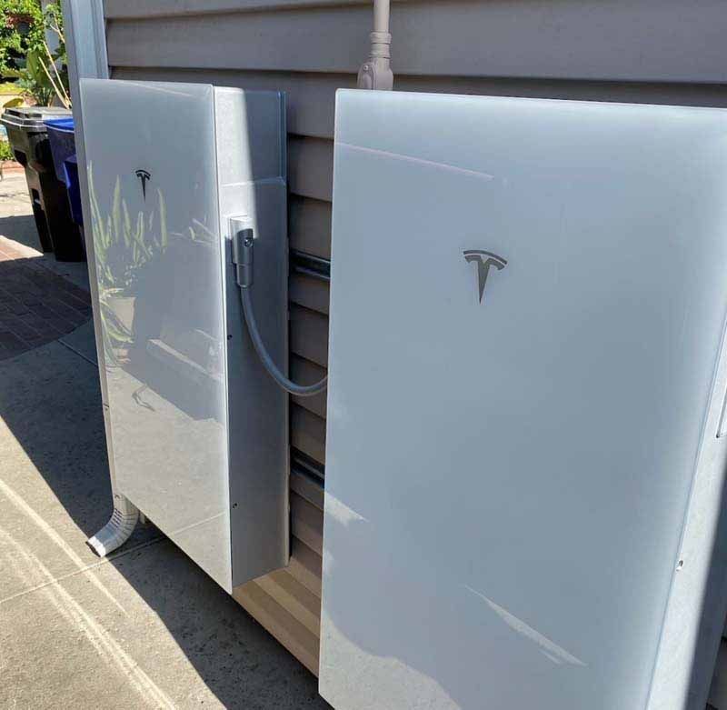 Two Tesla wall-mounted battery units beside a house exterior, one open showing wiring.