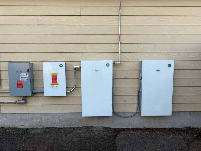 Four outdoor electrical panels mounted on a beige wall, with gray and white boxes connected by conduit.