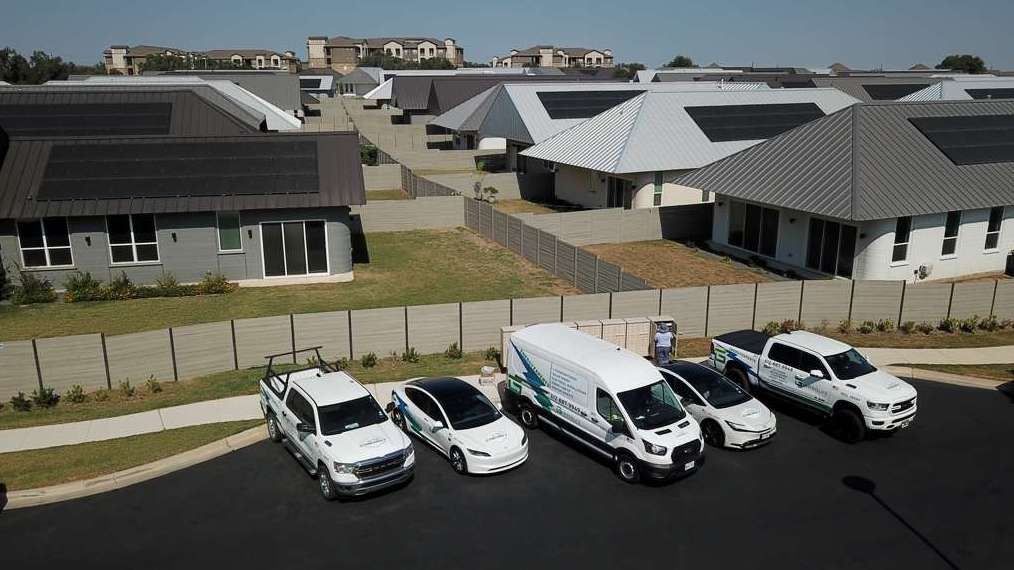 Several white vehicles parked along a residential street beside modern houses and a fenced lawn.