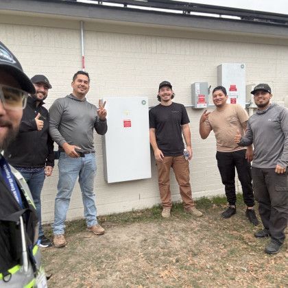 Five people posing outdoors beside a wall-mounted electrical box, several giving thumbs-up.