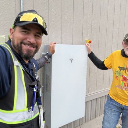 Two workers holding a white Tesla panel outdoors, one in a reflective vest and cap, the other in a yellow shirt