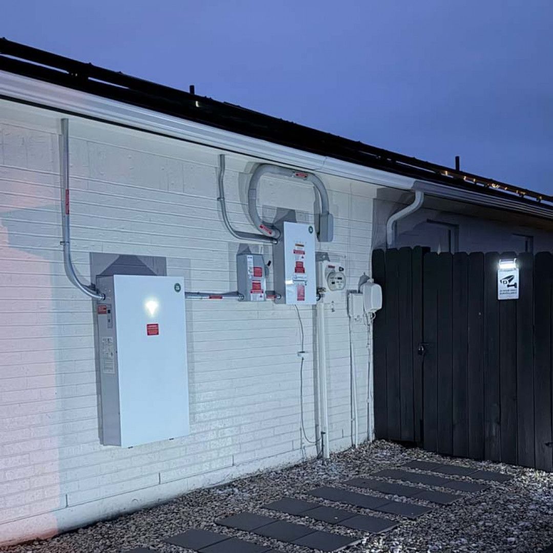 Exterior of a white brick building with utility boxes and meters beside a dark fence at dusk