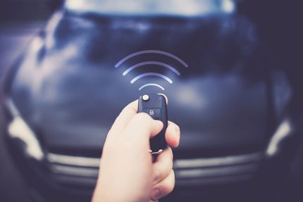 Una mano impugna un telecomando per auto verso un'auto scura, con un'icona digitale sovrapposta che indica una connessione wireless.