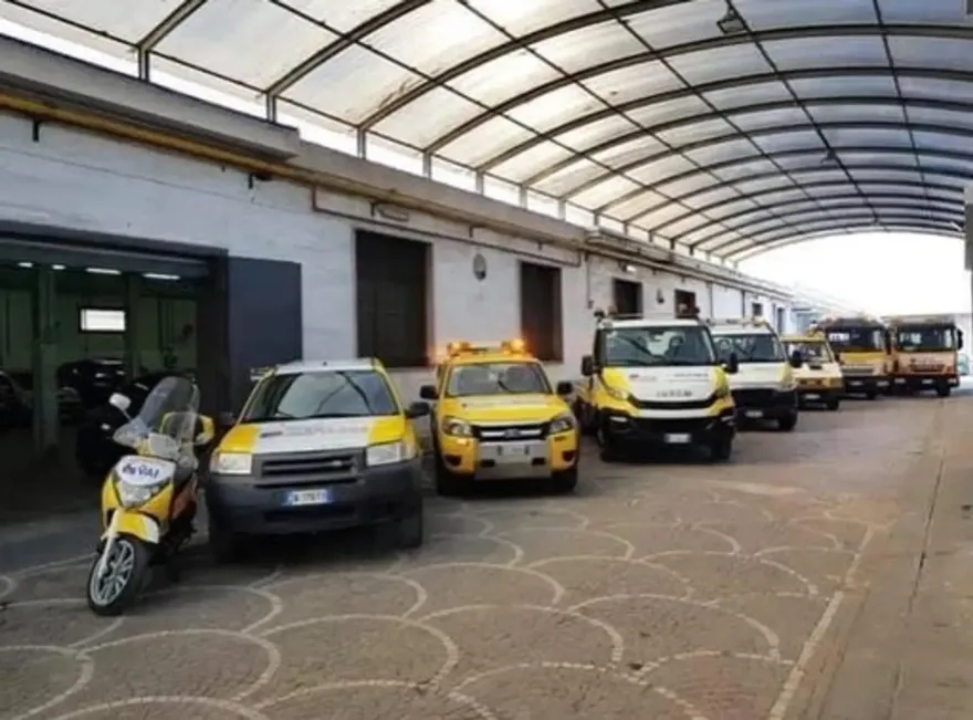 Una fila di veicoli di servizio gialli e bianchi e uno scooter parcheggiati sotto una tettoia esterna.