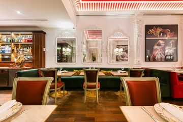 Sala de jantar elegante de restaurante com bancos estofados verdes, cadeiras com detalhes em vermelho e obras de arte emolduradas em paredes brancas.