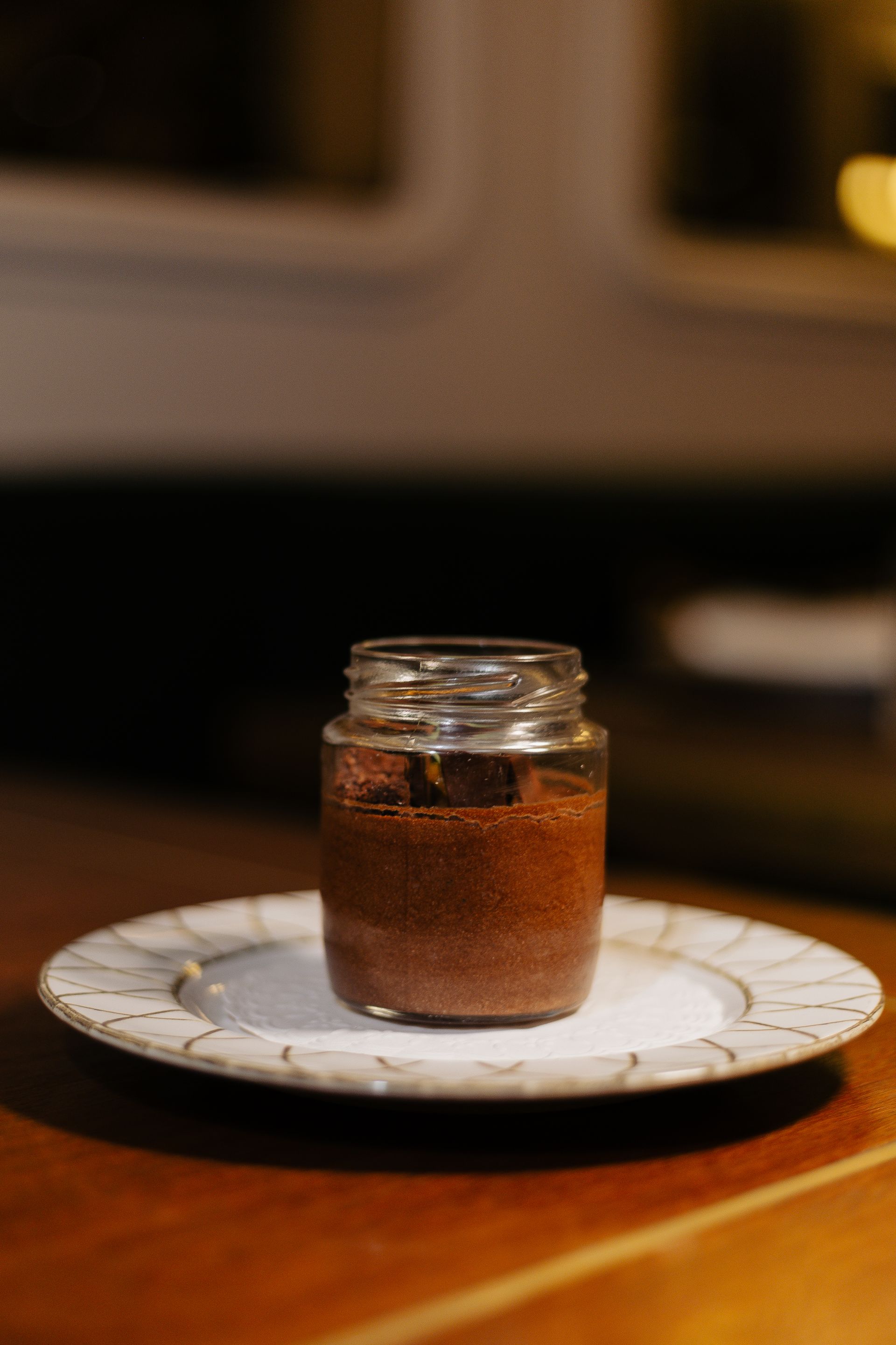 Pequeno pote de vidro contendo uma sobremesa escura sobre um prato branco em uma mesa de madeira.