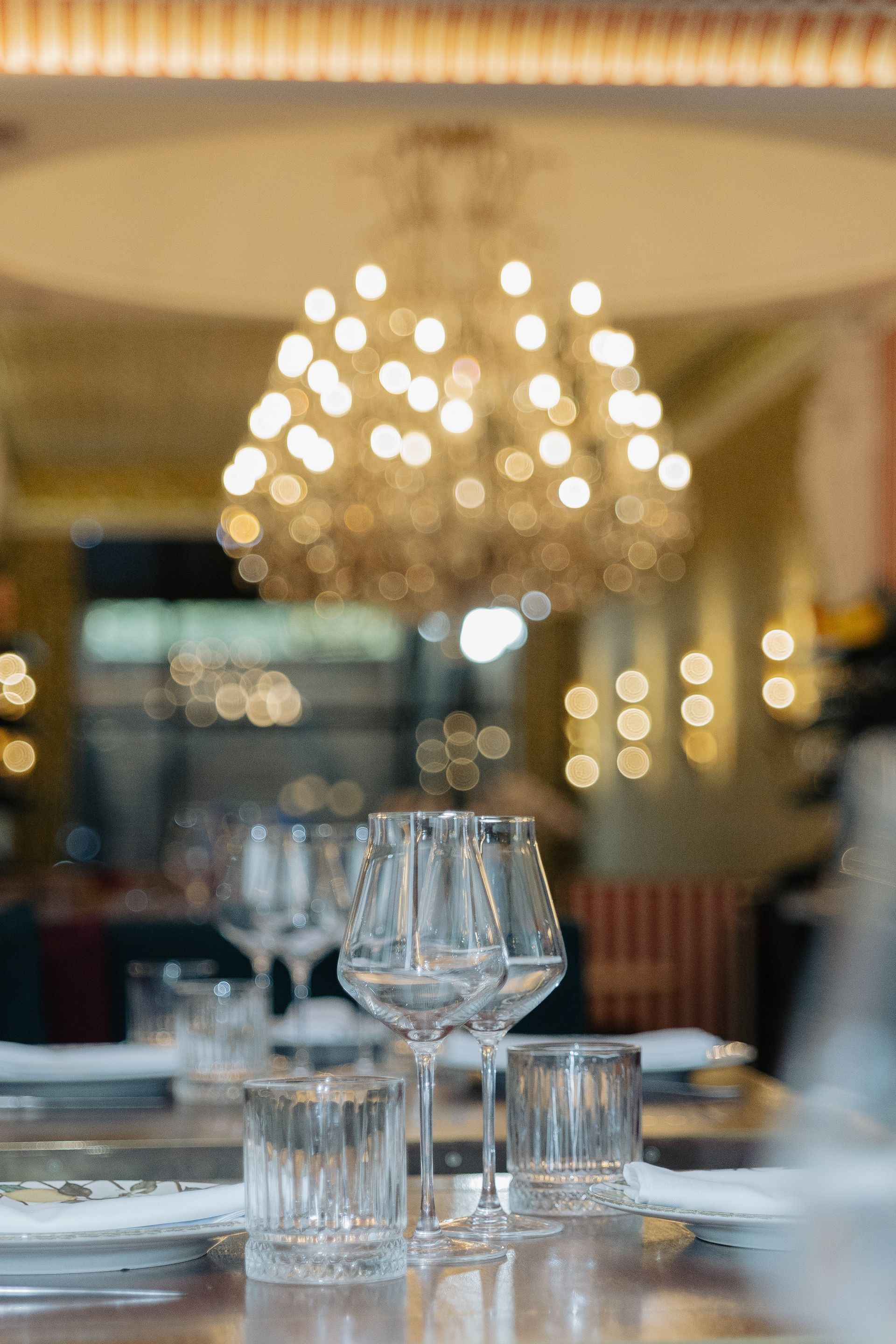 Mesa de restaurante elegante com taças de vinho e lustre à luz de velas ao fundo.