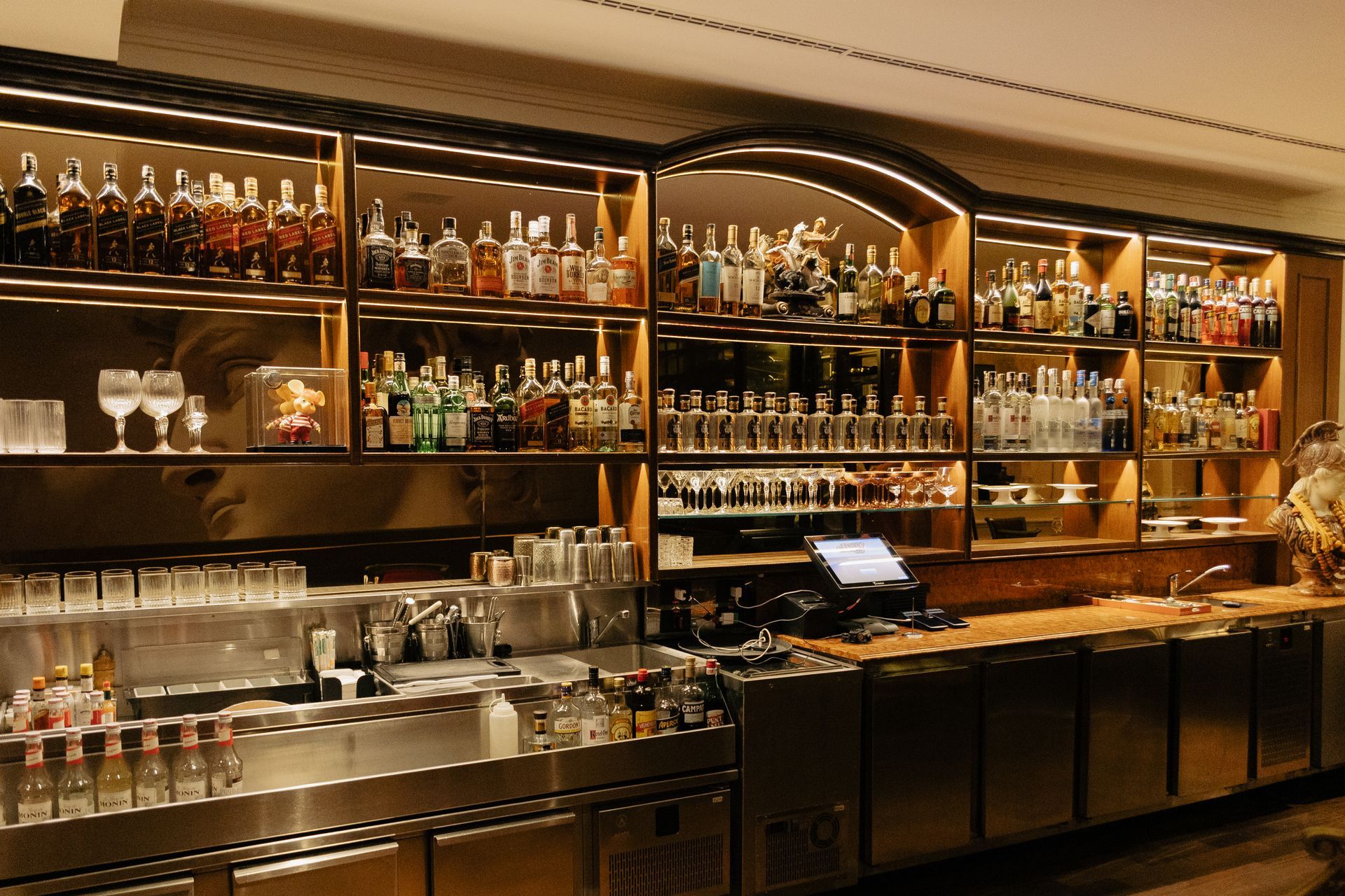 Bar com iluminação aconchegante, prateleiras repletas de garrafas de bebidas, copos e um balcão comprido.