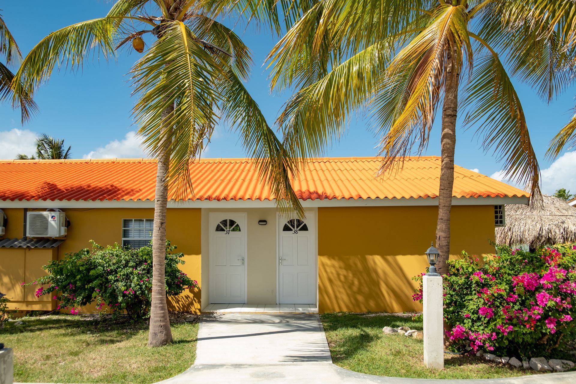 Yellow building with orange roof, white doors, palm trees, and green bushes.