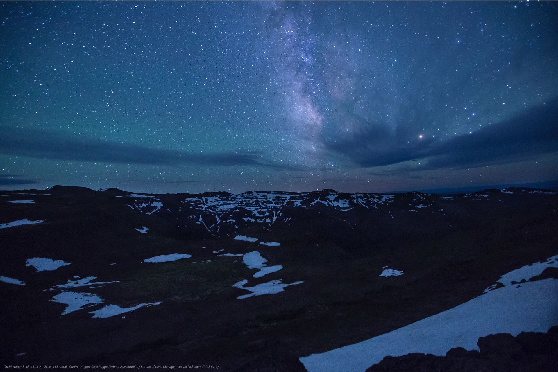Night sky with Milky Way over snow-patched terrain, subtle aurora borealis.