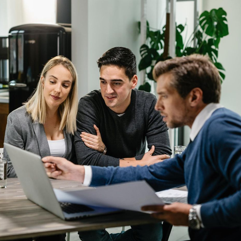 Three coworkers discuss a laptop at a table in a bright office