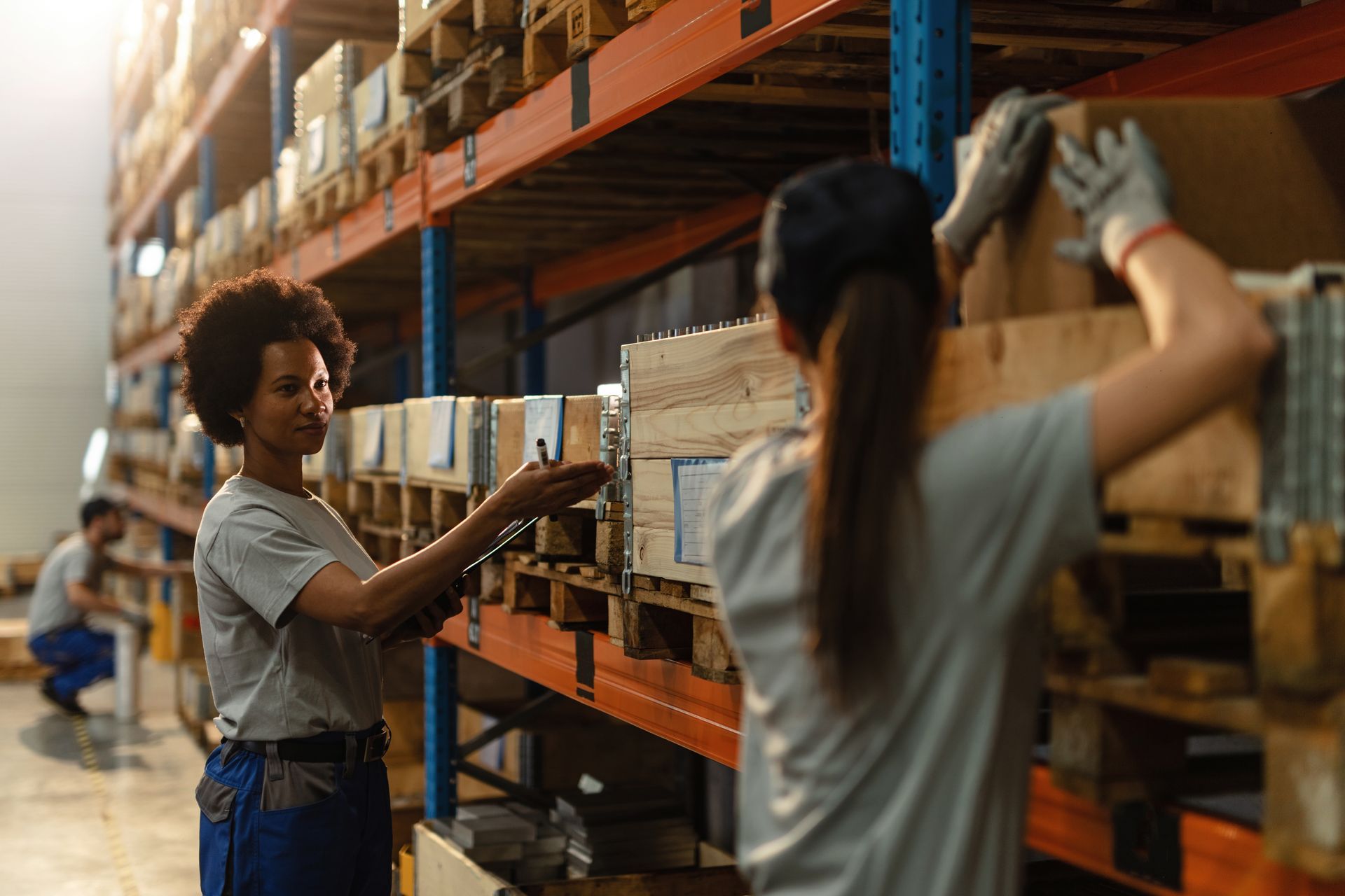 Workers stocking boxes on warehouse shelves with orange racks and a hand truck in the aisle