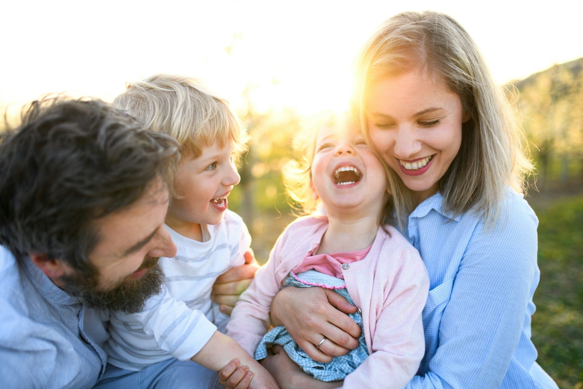 Family laughing together outdoors in warm sunlight, with two adults and two children close together.