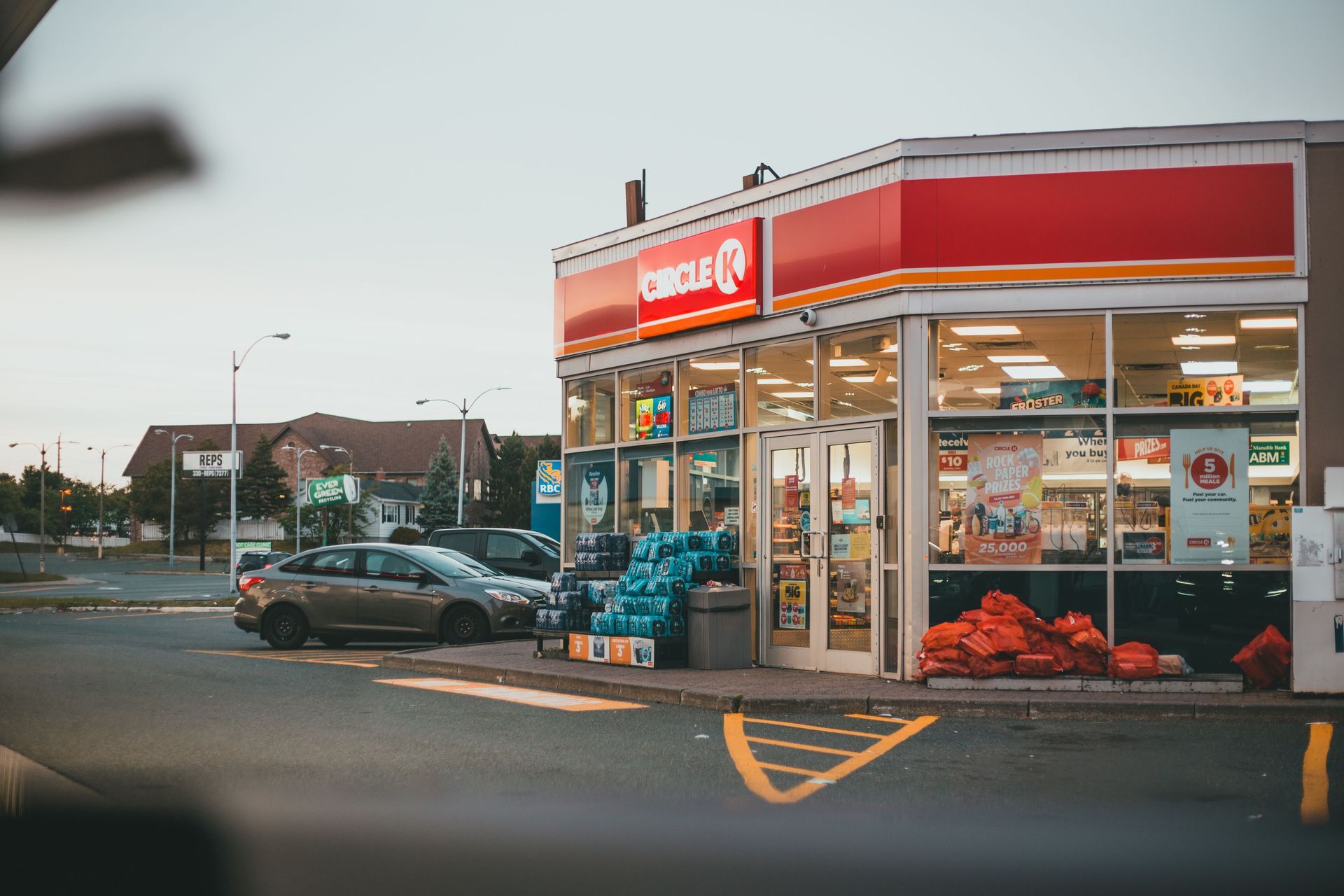 Corner grocery store at dusk with red sign, parked cars, and produce stacked outside the entrance.