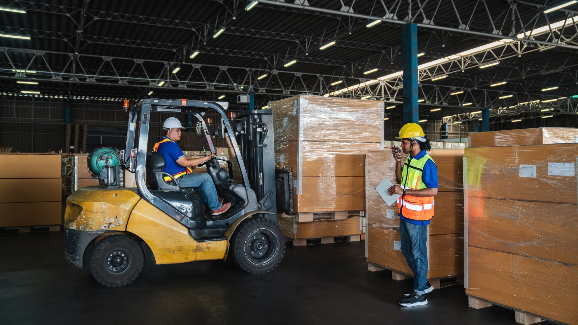 Forklift operator moving wrapped pallets in a warehouse while a worker in a safety vest monitors nearby