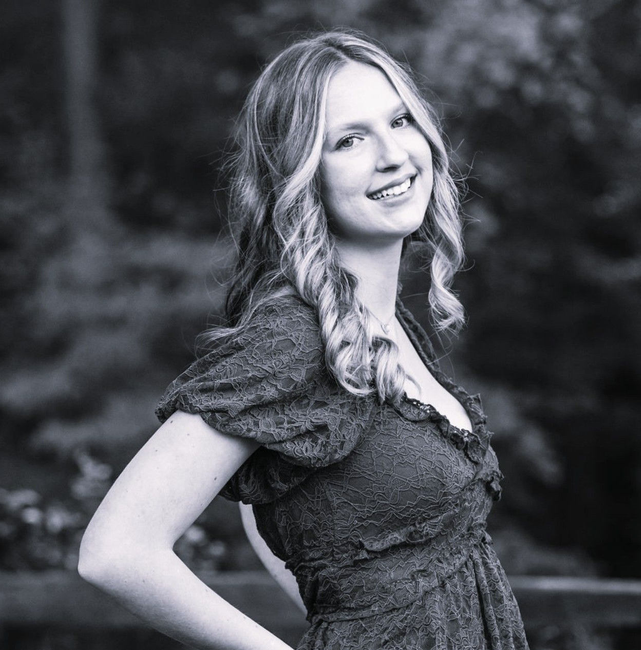 Smiling person in a dark dress standing outdoors with hands on hips, in black-and-white.