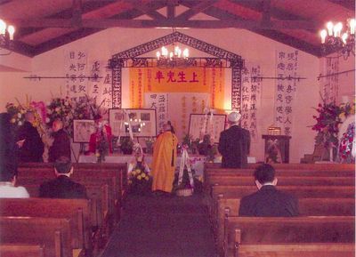 Funeral service in an Asian-style chapel. People seated, monk in orange robe, altar with banners and floral arrangements.