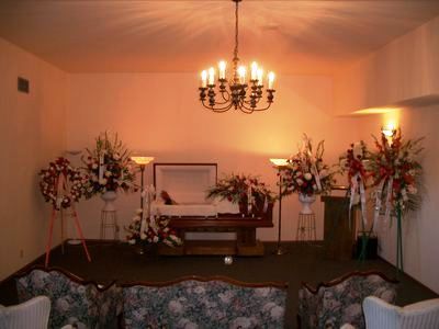 Funeral home interior with open casket, floral arrangements, and chandelier.