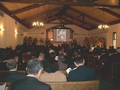 Funeral service: People seated in a chapel, looking at a screen displaying a photo. Flowers and chandeliers decorate the space.