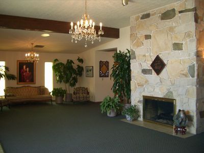 Interior of a building with a stone fireplace, chandeliers, plants, a sofa, and dark carpet.