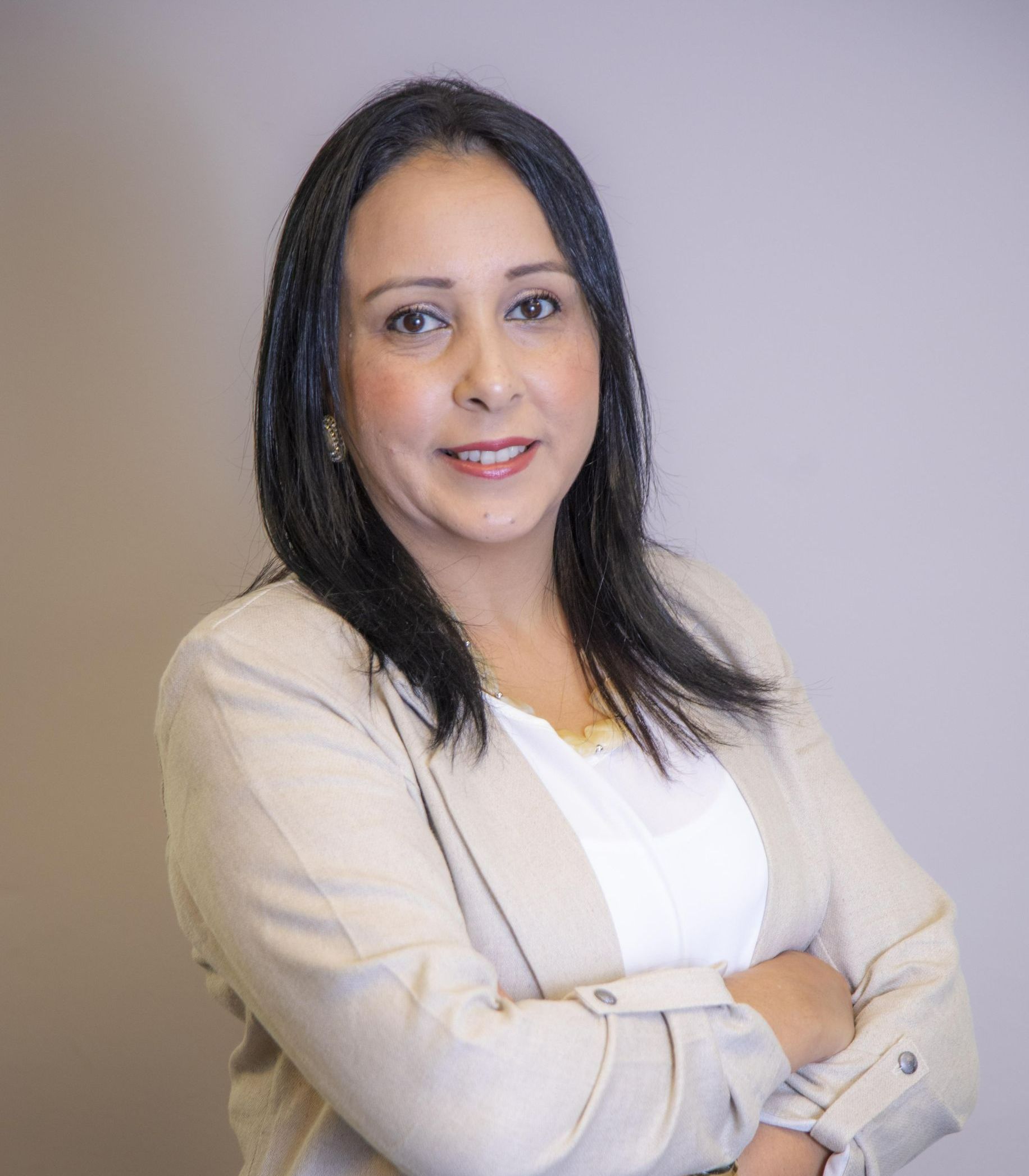 Retrato de uma mulher sorridente com os braços cruzados, vestindo um blazer claro contra um fundo neutro.