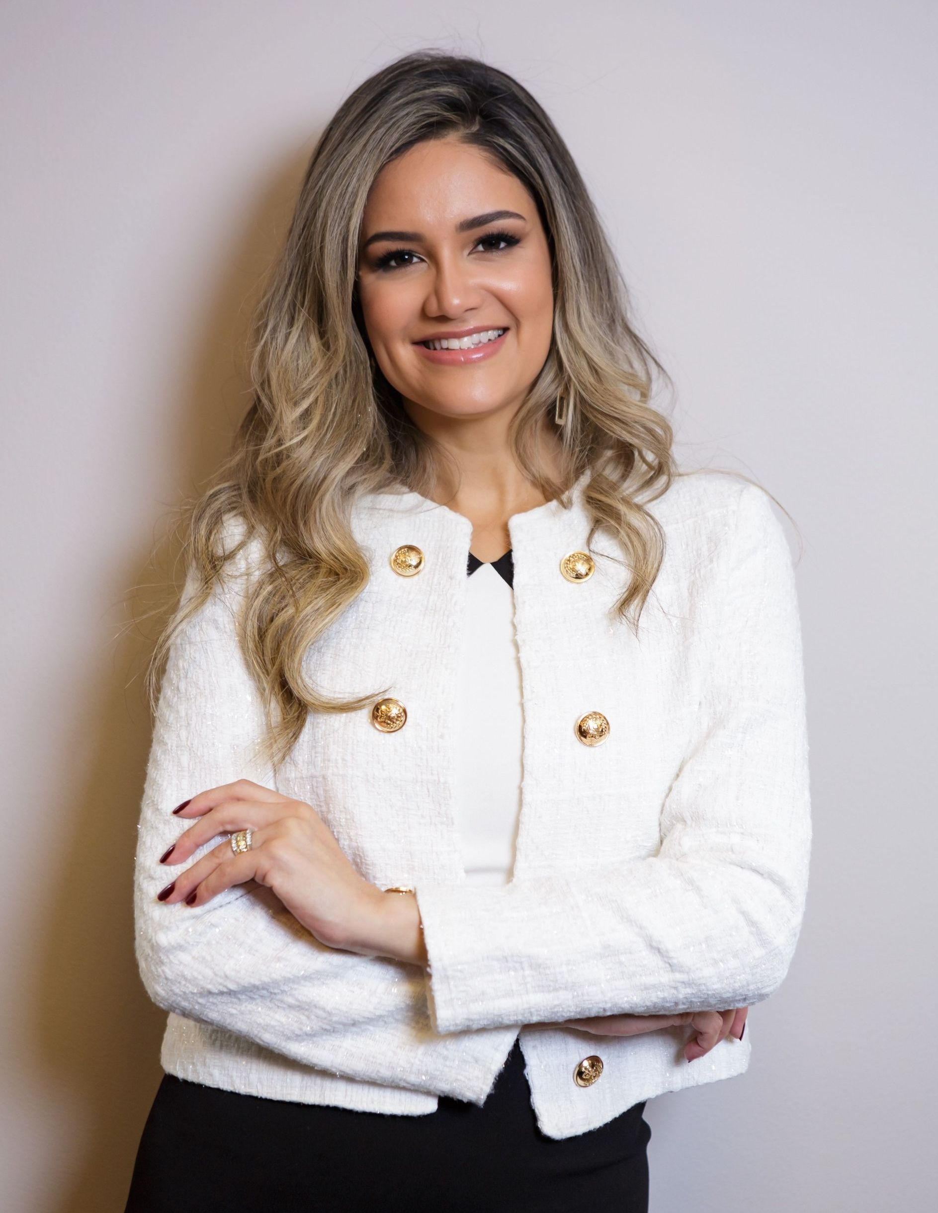 Mulher sorridente de jaqueta branca com botões dourados, em pé com os braços cruzados contra um fundo cinza.