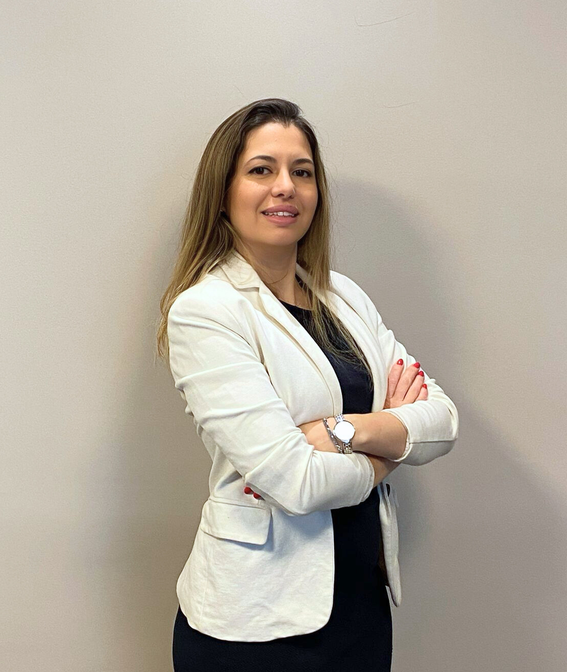 Pessoa vestindo blazer branco e vestido preto, em pé com os braços cruzados contra uma parede lisa.