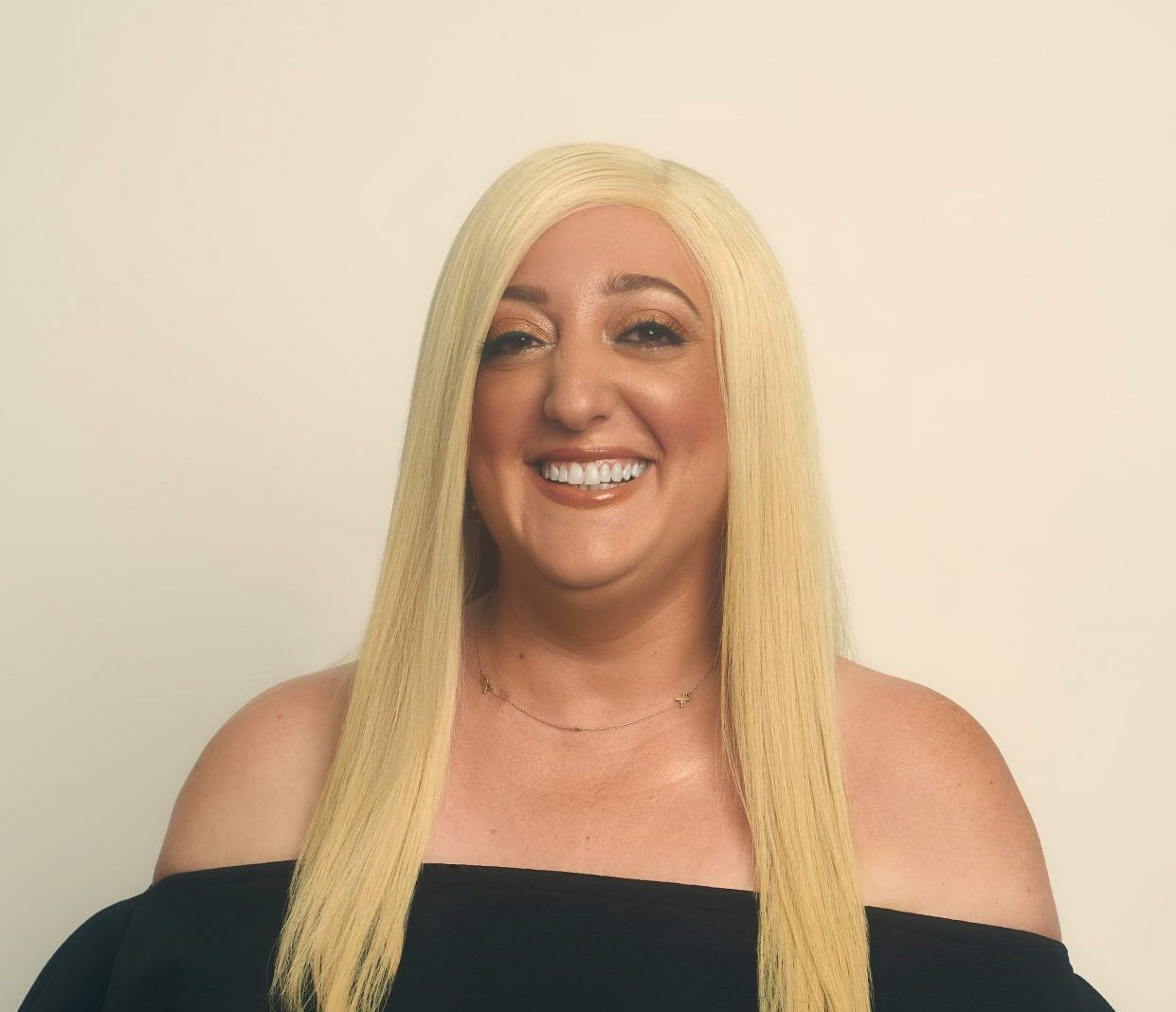 Woman with long, blonde, crimped hair wearing a black off-the-shoulder top smiles at the camera.