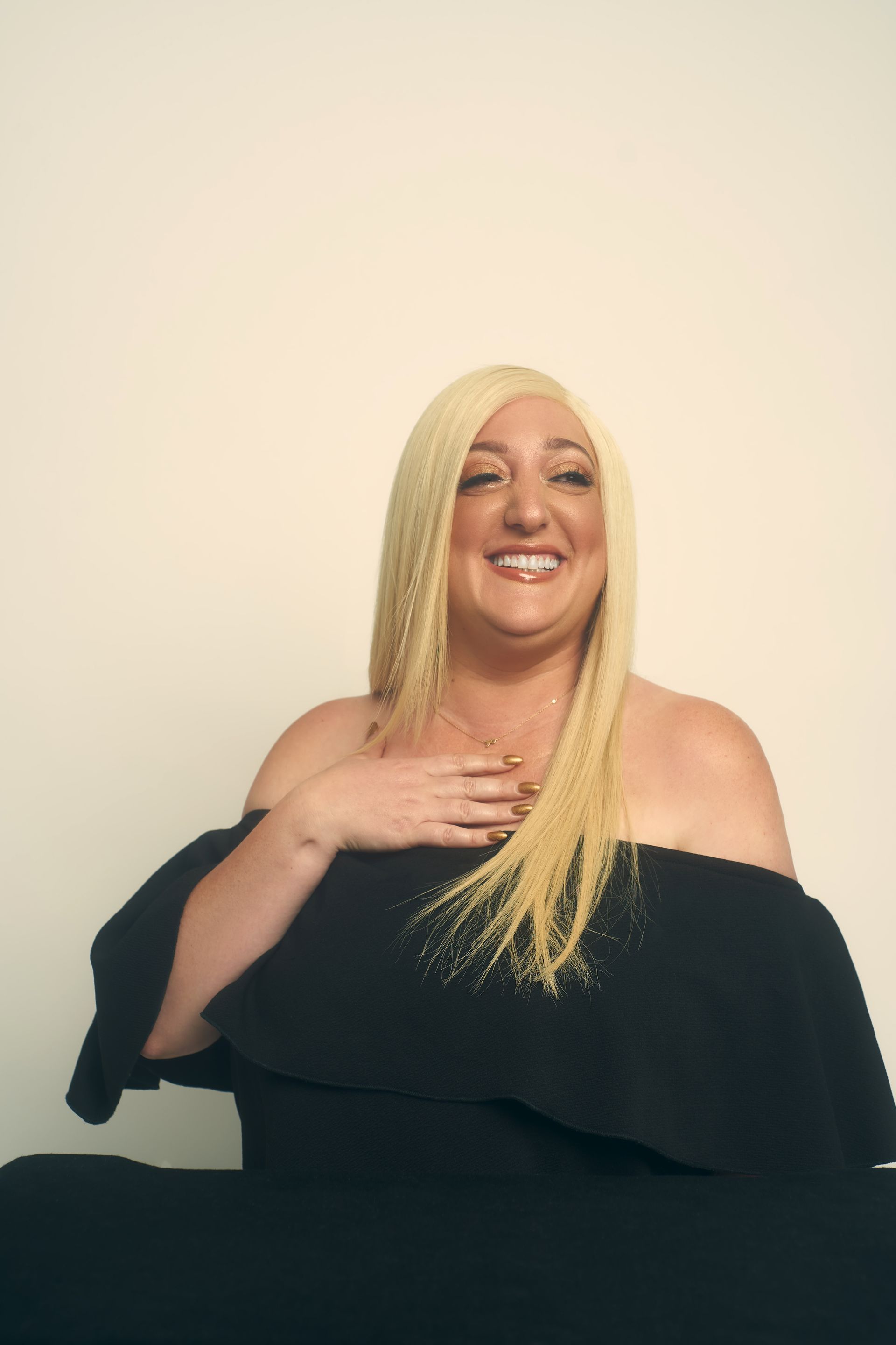 Woman with blonde hair, in a black off-the-shoulder dress, smiling and posing in a studio.