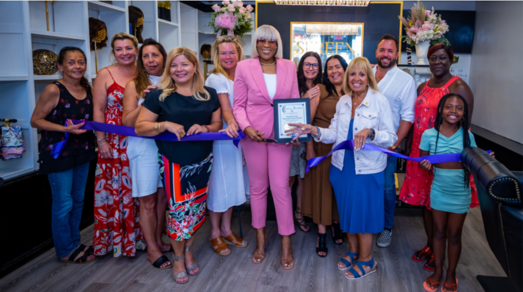 Group of people cutting a purple ribbon at a salon opening; a woman in pink holds a certificate.