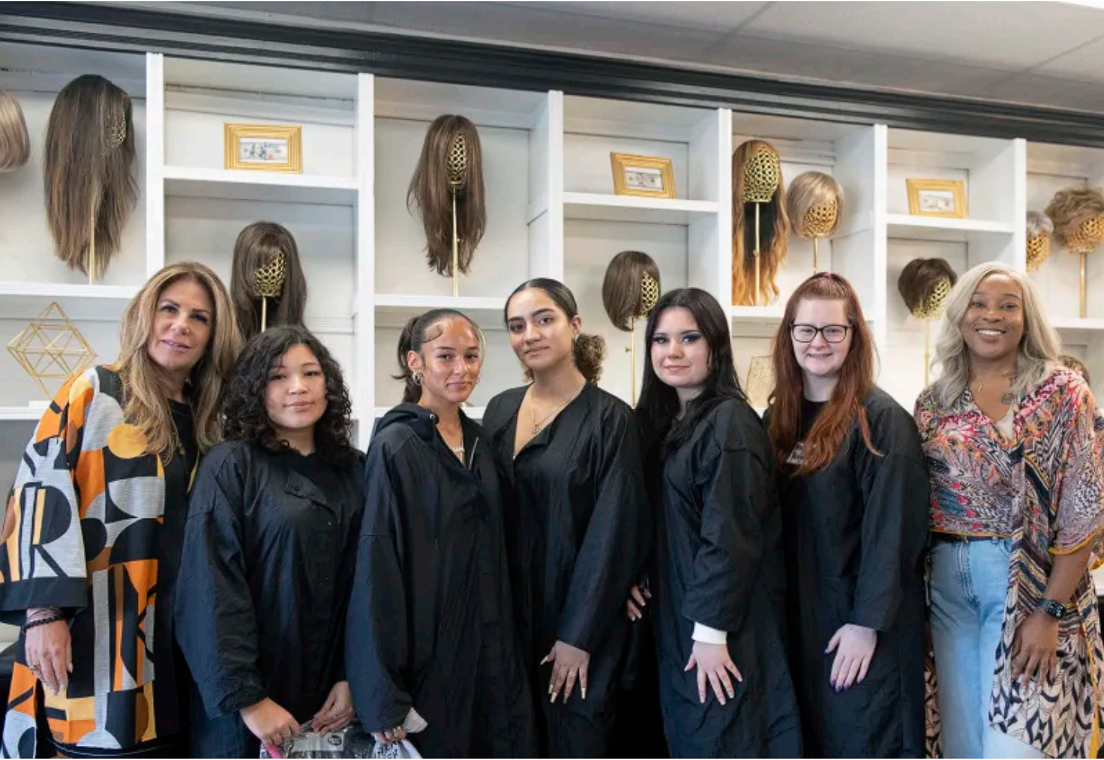 Group of women in black robes pose in front of a display of wigs.