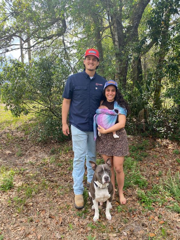 Man in red cap, woman with a baby, and adog posing outdoors among trees and leaves