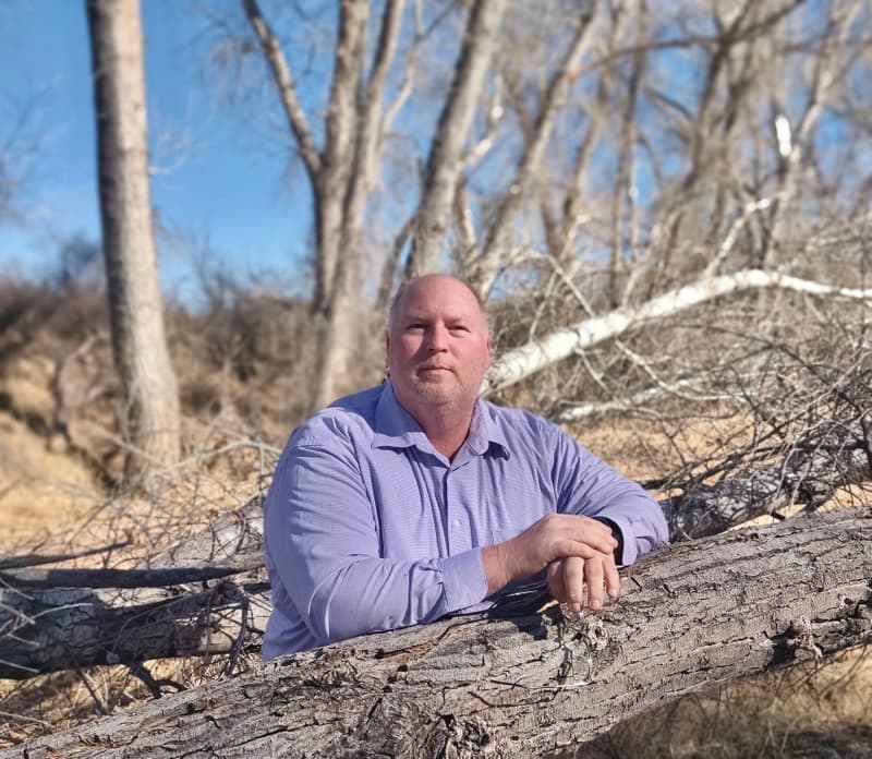 Man in a lavender shirt leaning on a fallen tree in a wooded area under a blue sky