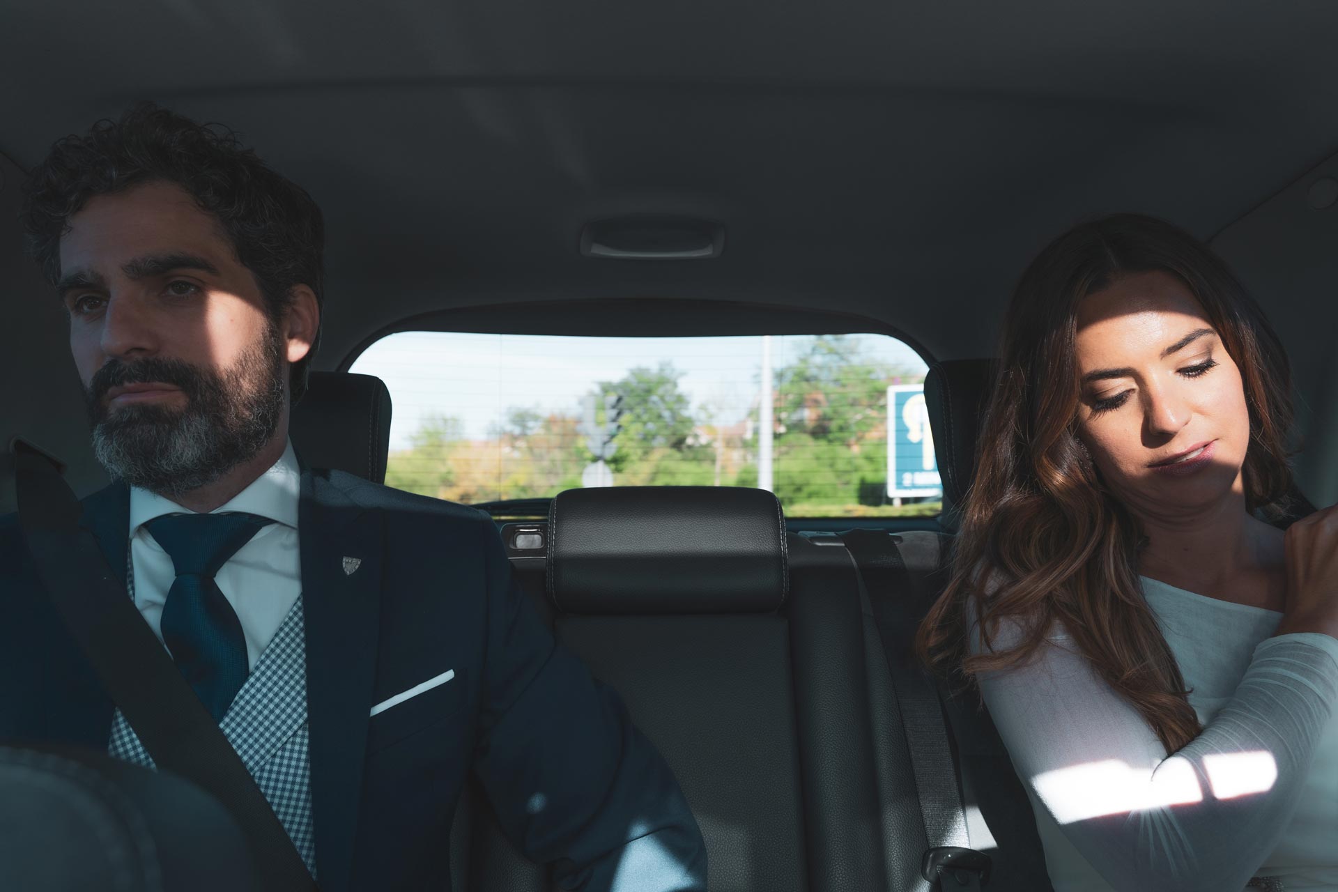 Two passengers sit in the back seat of a car, one looking ahead and the other resting with eyes closed.