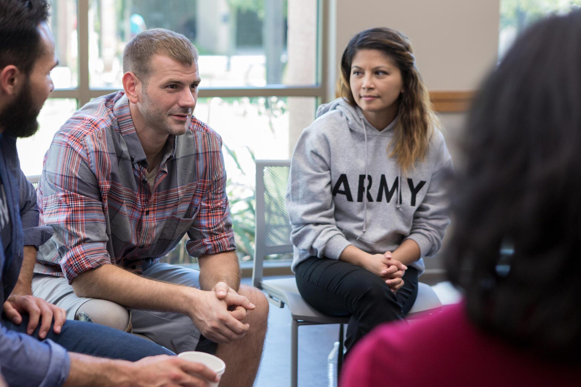 Small group in discussion, seated indoors; one person in a gray ARMY sweatshirt listens attentively.