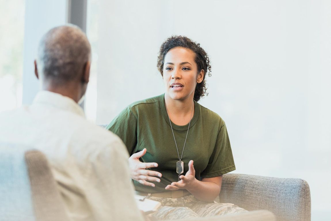 Two people in a bright room talking across a table, one gesturing while holding a mug.