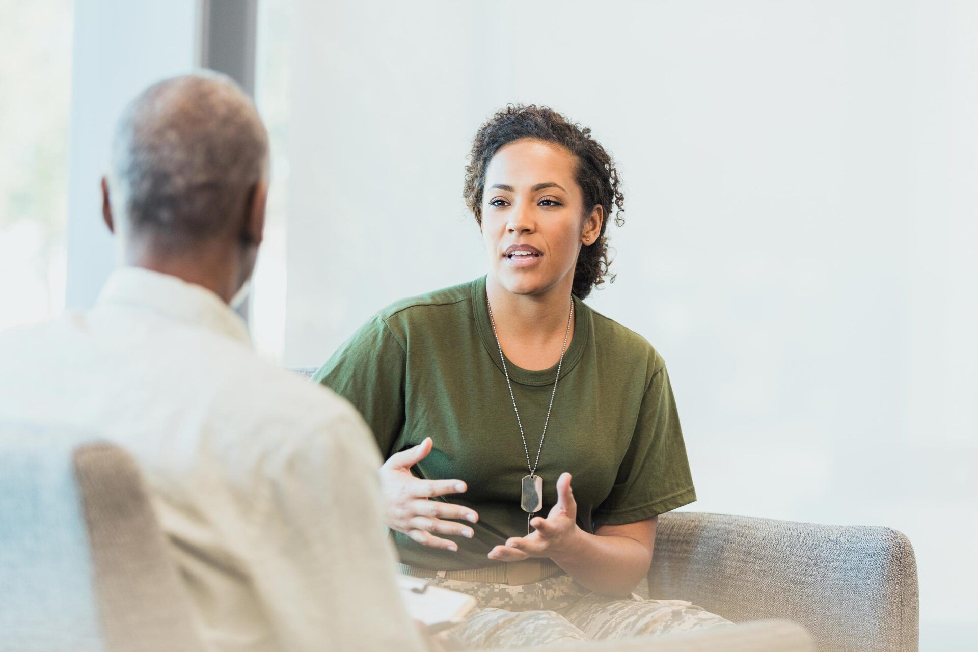 Two people in a bright room talking across a table, one gesturing while holding a mug.