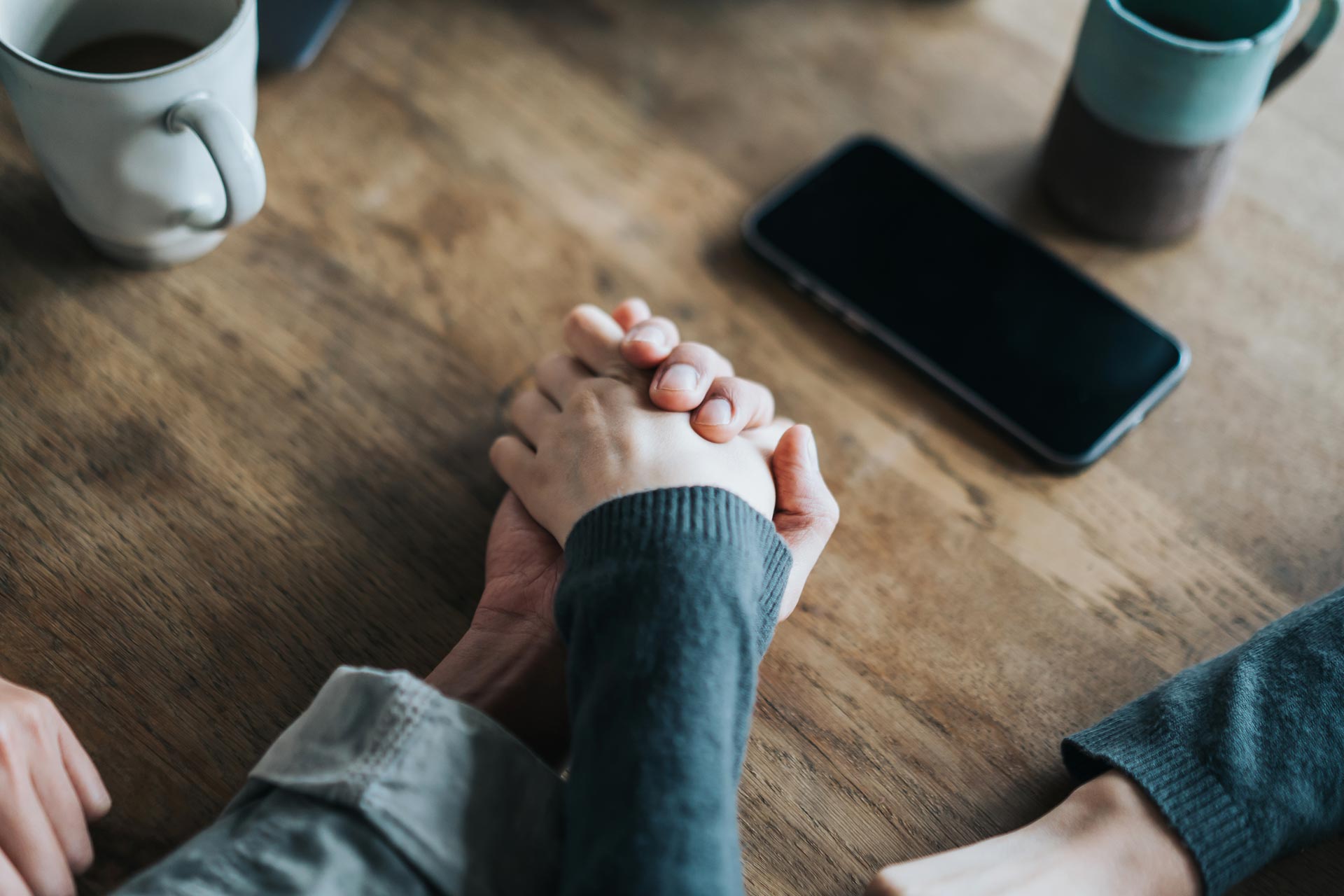 Two people clasping hands across a wooden table with coffee mugs and a smartphone nearby.