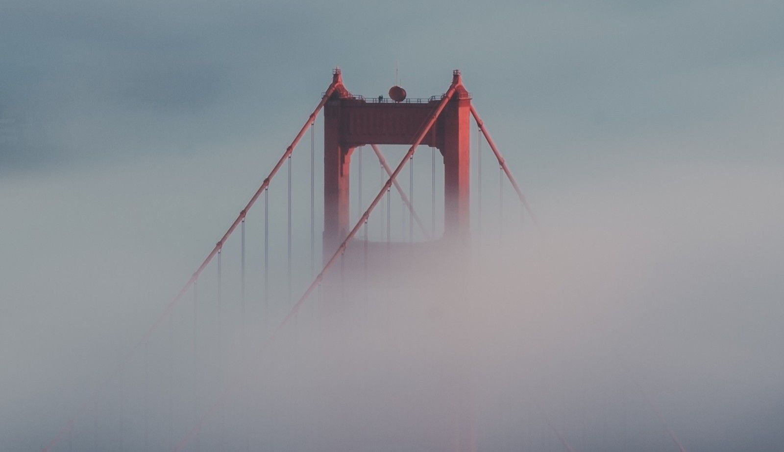 Red suspension bridge tower rising above thick fog at dawn