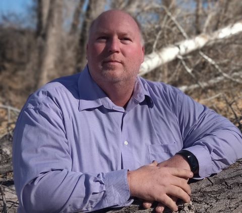 Man in a lavender shirt leaning on a rock outdoors with blurred trees in the background
