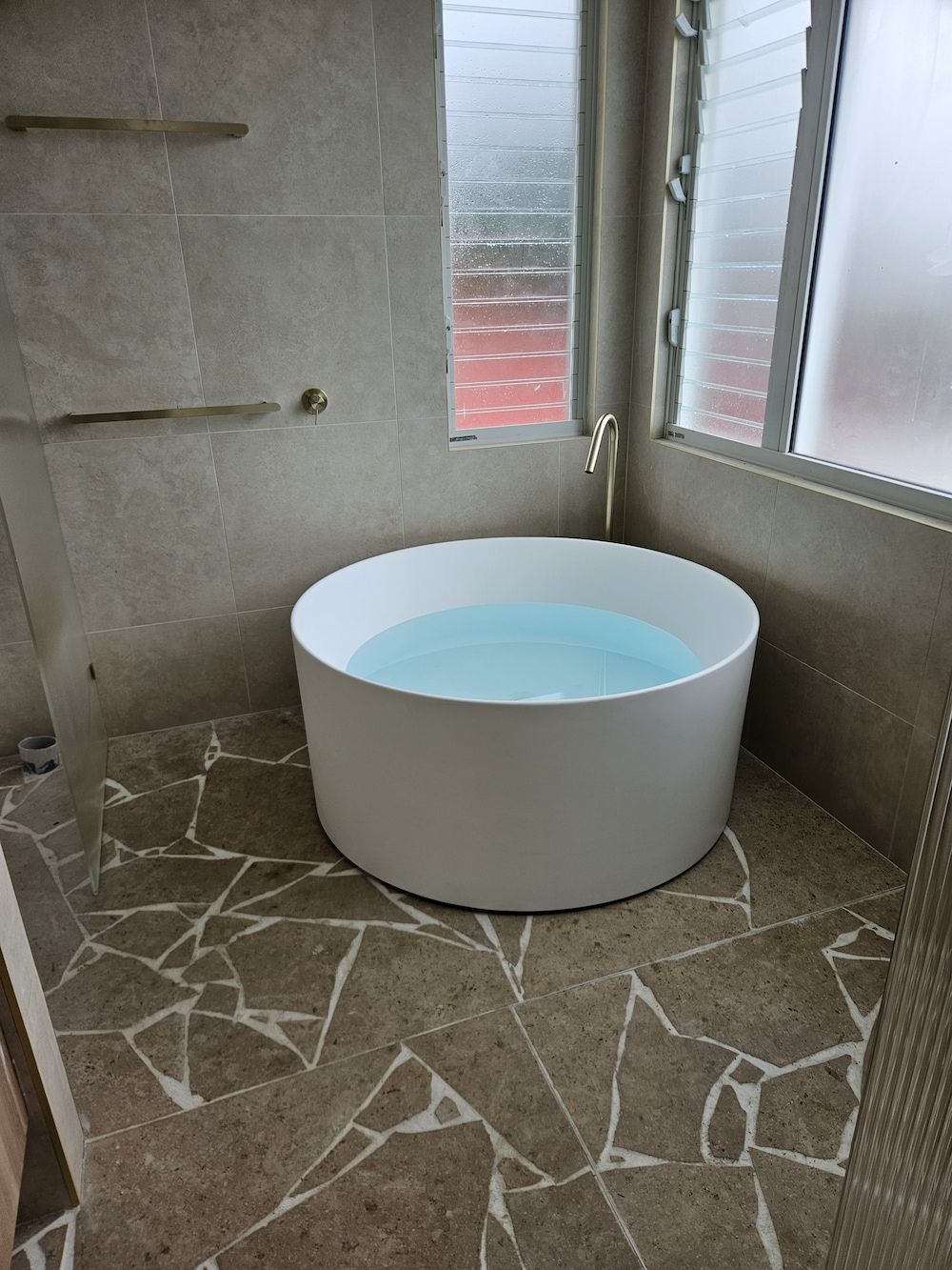 Circular white bathtub filled with water, set in a bathroom with mosaic tile floor, next to a window. — Kolora Projects in Robina, QLD